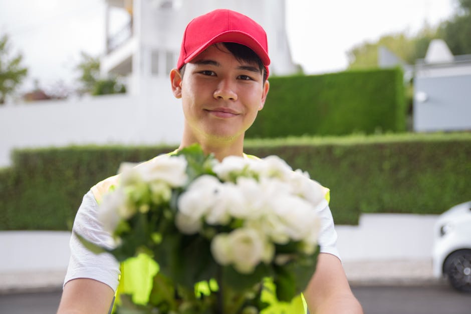 A cheerful delivery person dressed in a high-visibility yellow vest and a red cap, smiling as he extends a large bouquet of white flowers towards the camera. The bouquet features a mix of fresh, fully