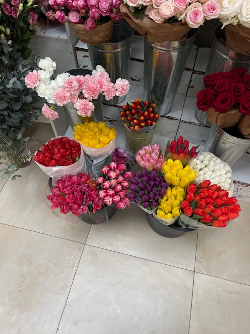 A vibrant display of fresh flowers arranged in various bouquets and containers at a florist shop. The image features pink and white carnations, red roses, tulips in shades of pink, white, yellow, and 