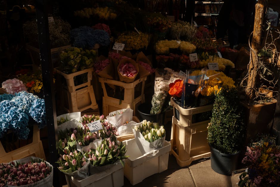 An outdoor flower market stall displaying a variety of fresh floral arrangements and bouquets. In the foreground, there are white and pink tulips wrapped in white paper, alongside pink and purple hyac