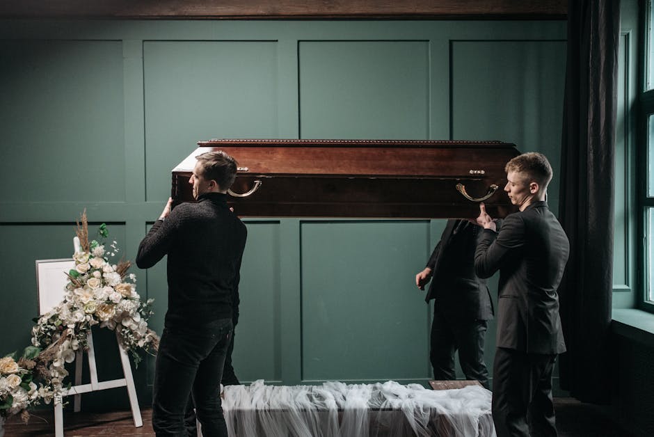 Two young men dressed in black suits are carrying a closed wooden casket into a room with dark green walls and large windows on the right. To the left, there is a floral arrangement featuring white ro