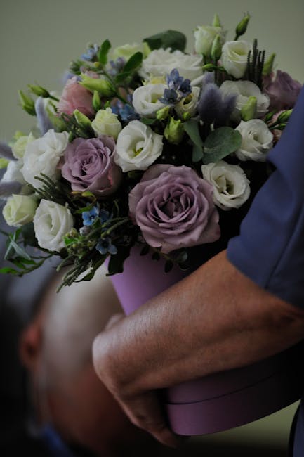 A floral bouquet featuring a mix of cream and soft purple roses, accented with delicate blue flowers and lush green foliage, arranged in a rounded style and held by a person's hand. The bouquet is wra