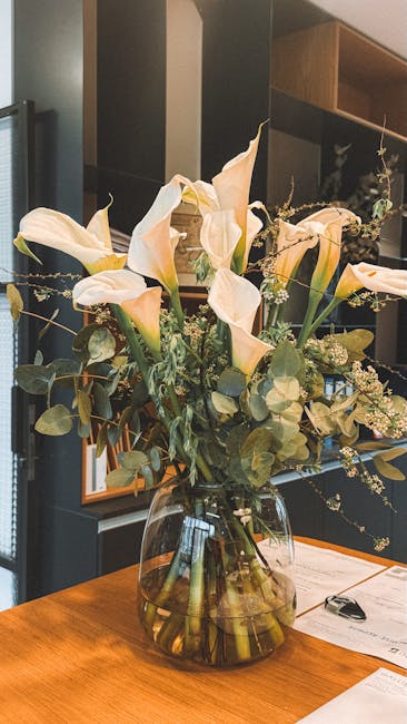 A floral arrangement of white calla lilies with smooth, elegant petals and long, slender stems, complemented by sprigs of eucalyptus with round, silver-green leaves, all arranged in a clear, round gla