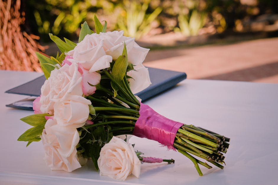 A bouquet of fresh white and pale pink roses arranged in a rounded, elegant style, with green leaves interspersed among the flowers. The stems are wrapped with a pink satin ribbon, which is neatly tie