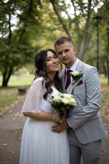 A newlywed couple stands outdoors in a park setting with lush green trees and a paved pathway. The bride has long, dark wavy hair and wears a flowing white wedding dress with delicate sheer sleeves. S